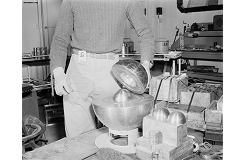 A photo of a man holding one half of a metal sphere to show a mockup of a sphere of plutonium encased within.